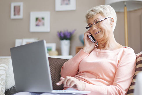 A senior woman makes a phone call while researching on her laptop.
