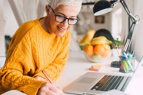 A senior woman writes notes down while working at a laptop.