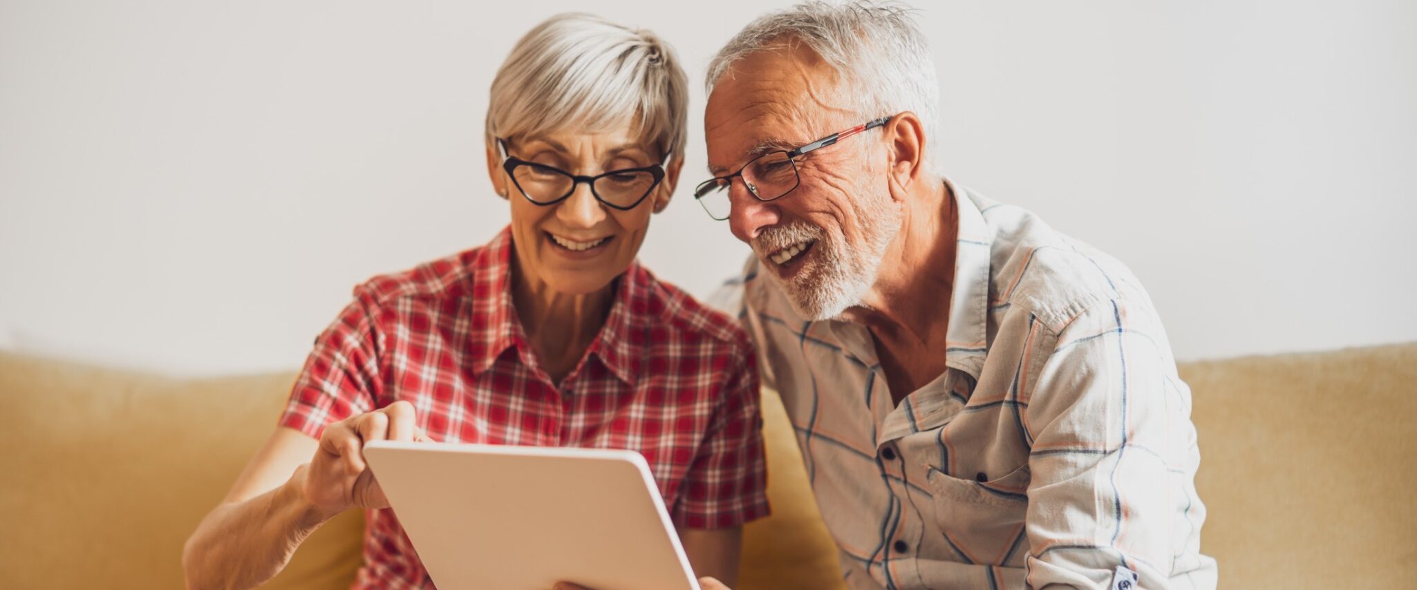 A smiling senior woman and man sit on a couch and look at a tablet together.