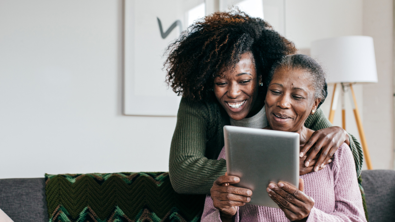 A woman leans over the couch to see the tablet screen her elderly mother is showing her